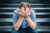 Outdoor portrait of sad young man sitting on stairs covering his face with hands.