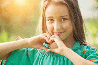 Young girl showing heart gesture and smiling outdoors.